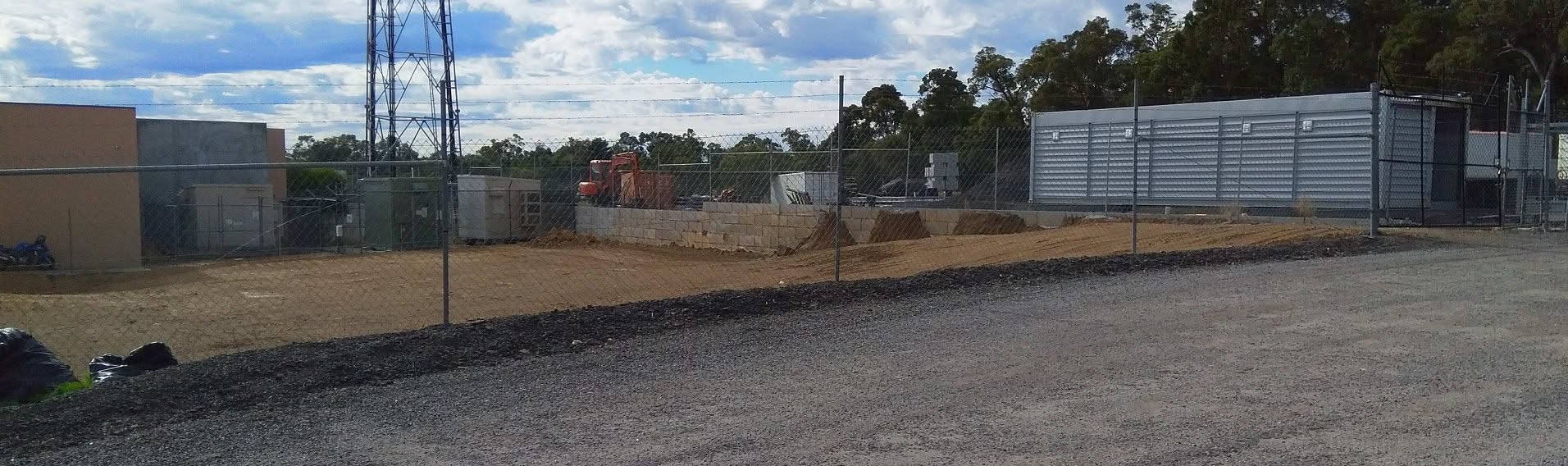 Fenced industrial yard with gravel hardstand and a storage building under a partly cloudy sky